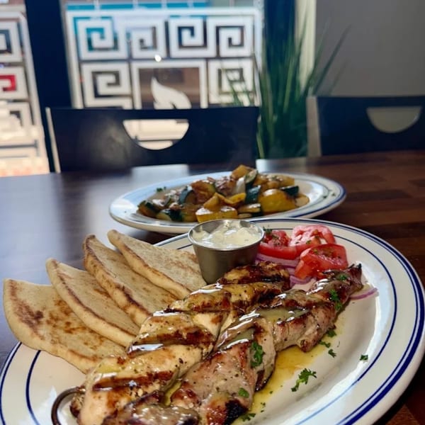 Grilled chicken skewers with pita bread, tzatziki, tomatoes, and a side of roasted vegetables.