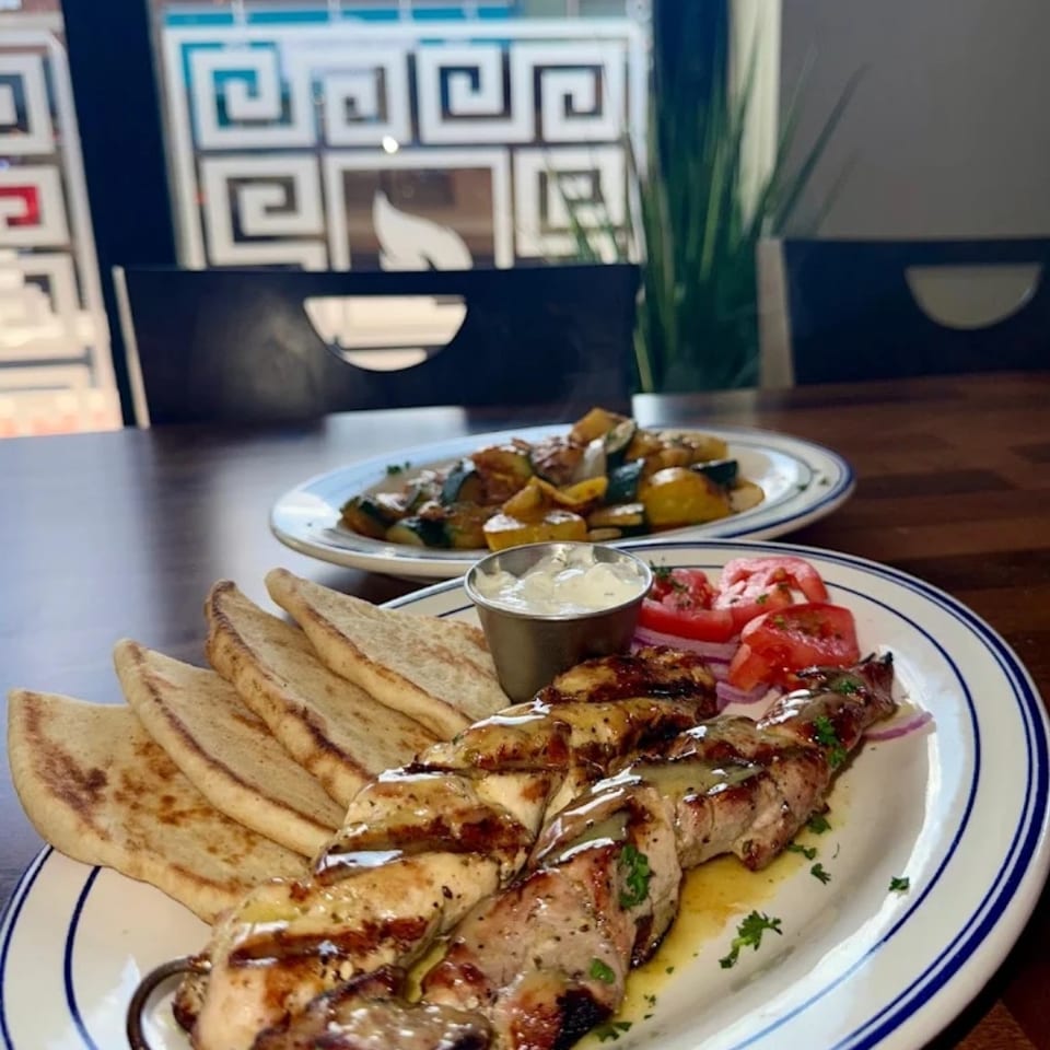 Grilled chicken skewers with pita bread, tzatziki, tomatoes, and a side of roasted vegetables.