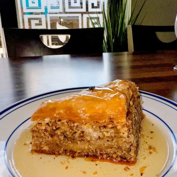 A slice of baklava with honey syrup on a white plate with blue trim, in a restaurant setting.