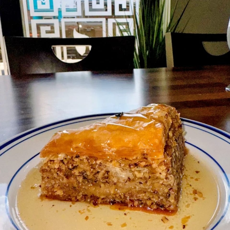 A slice of baklava with honey syrup on a white plate with blue trim, in a restaurant setting.